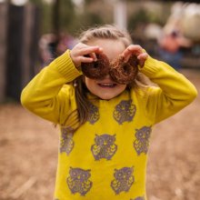 A little girl dressed in a fall sweater covers in owls holds 2 apple cider donuts over her eyes at Great Country Farms apple picking festival